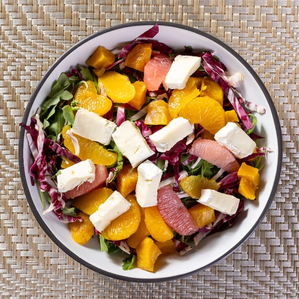 Overhead view of a colorful salad with citrus, cheese, and greens in a white bowl.