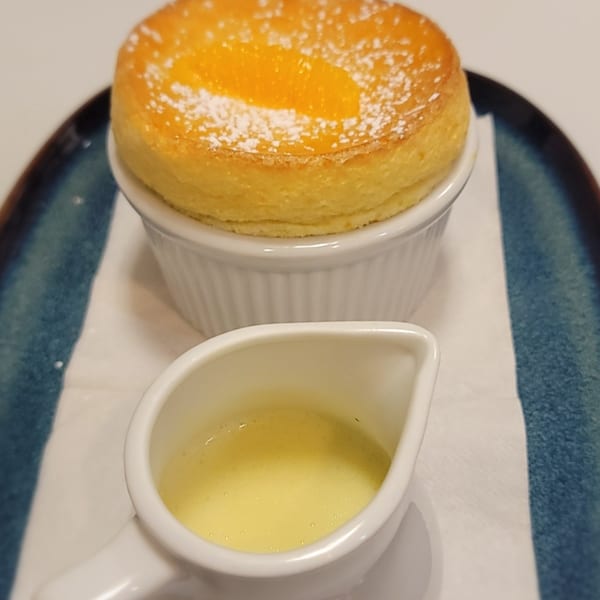 Orange souffle with powdered sugar in a white ramekin, served with a side of creme anglaise.