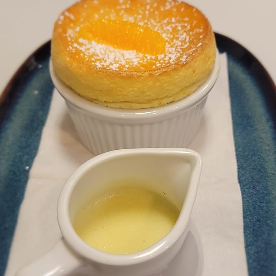Orange souffle with powdered sugar in a white ramekin, served with a side of creme anglaise.