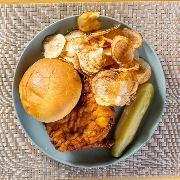 Fried chicken sandwich with a bun, potato chips, and a pickle spear on a blue plate.