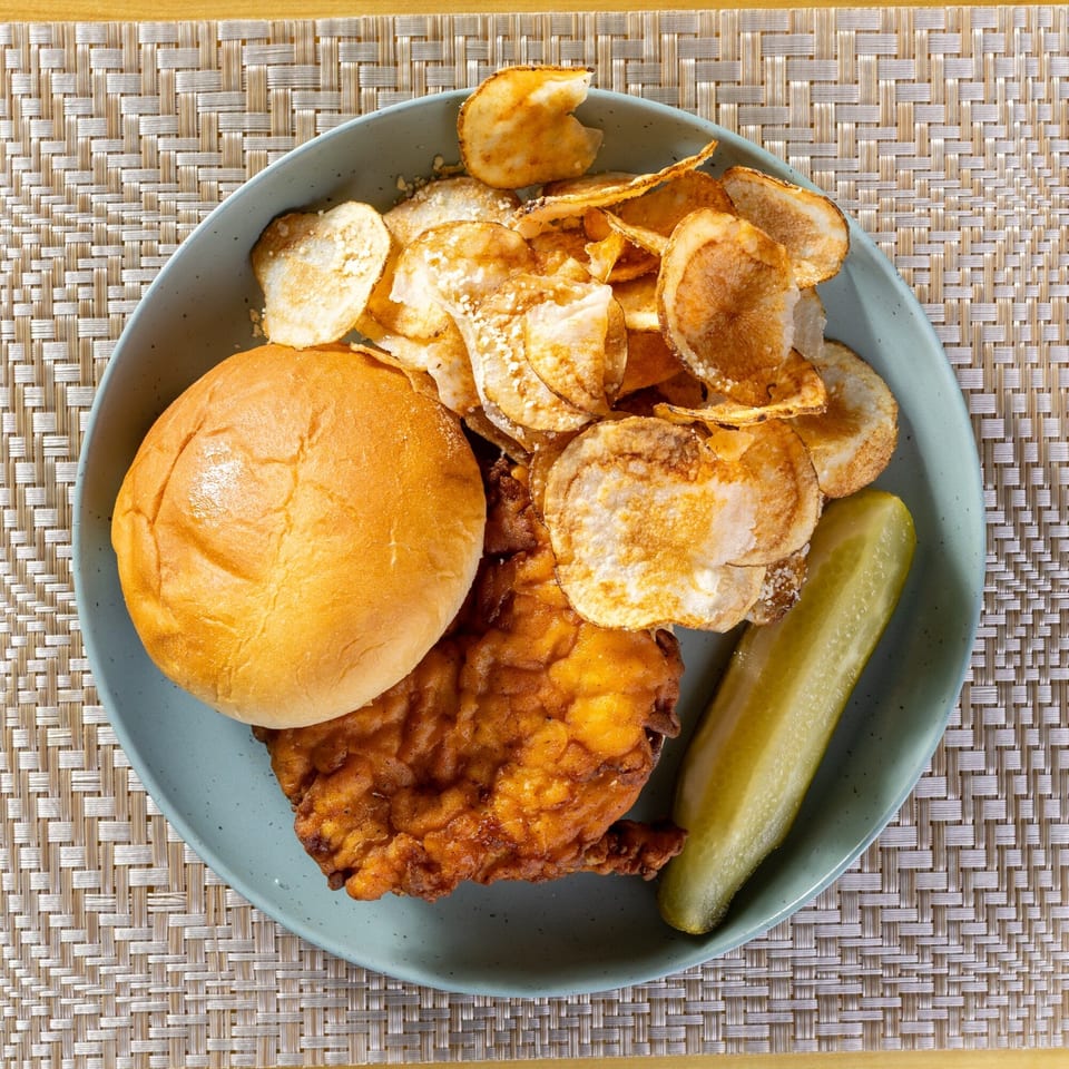 Fried chicken sandwich with a bun, potato chips, and a pickle spear on a blue plate.