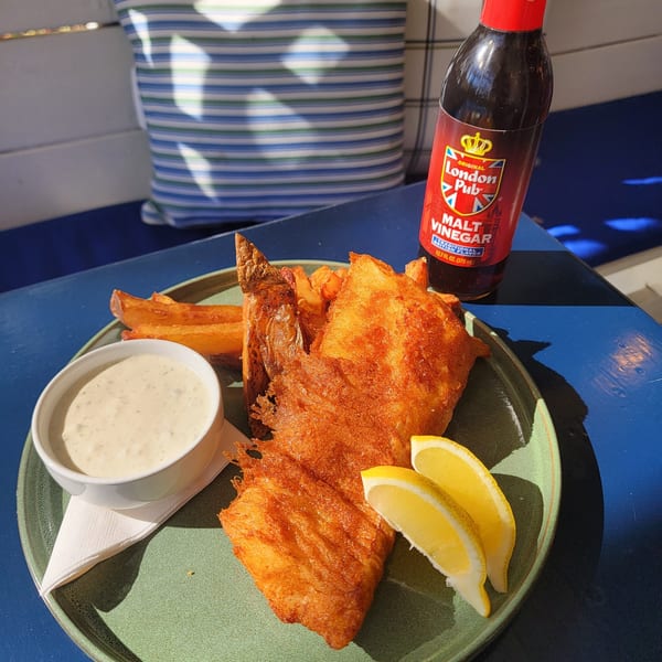 Golden-brown fish and chips with lemon wedges, tartar sauce, and malt vinegar on a blue table.