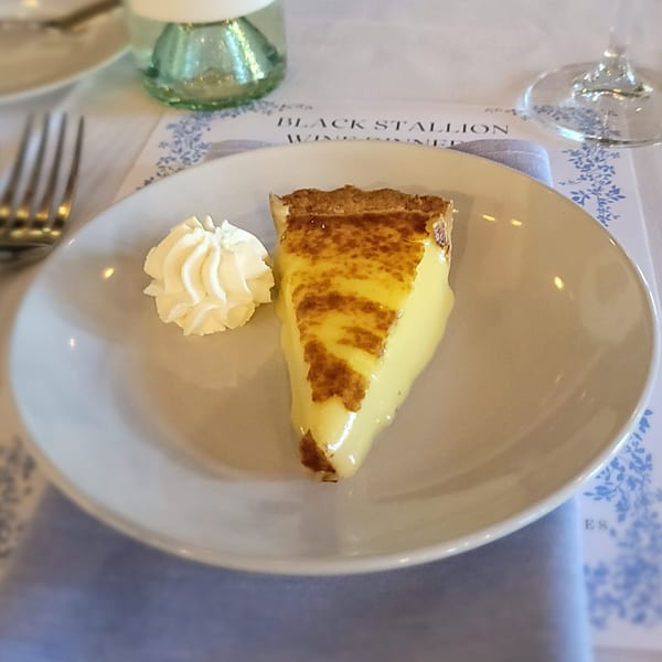 A slice of lemon tart with whipped cream on a white plate, set on a patterned placemat.