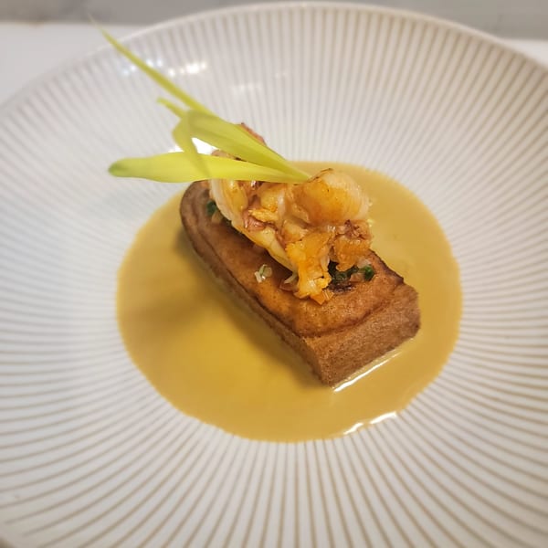 A rectangular piece of toast topped with shrimp and green garnish, in a yellow sauce on a white plate.