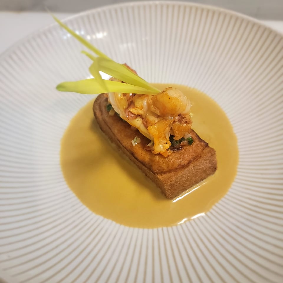 A rectangular piece of toast topped with shrimp and green garnish, in a yellow sauce on a white plate.