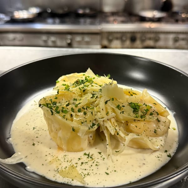 Creamy pasta dish with shaved cheese and fresh herbs in a black bowl, with a kitchen in the background.