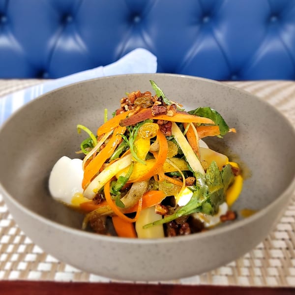 A vibrant salad with shaved carrots, greens, nuts, and golden raisins in a gray bowl.